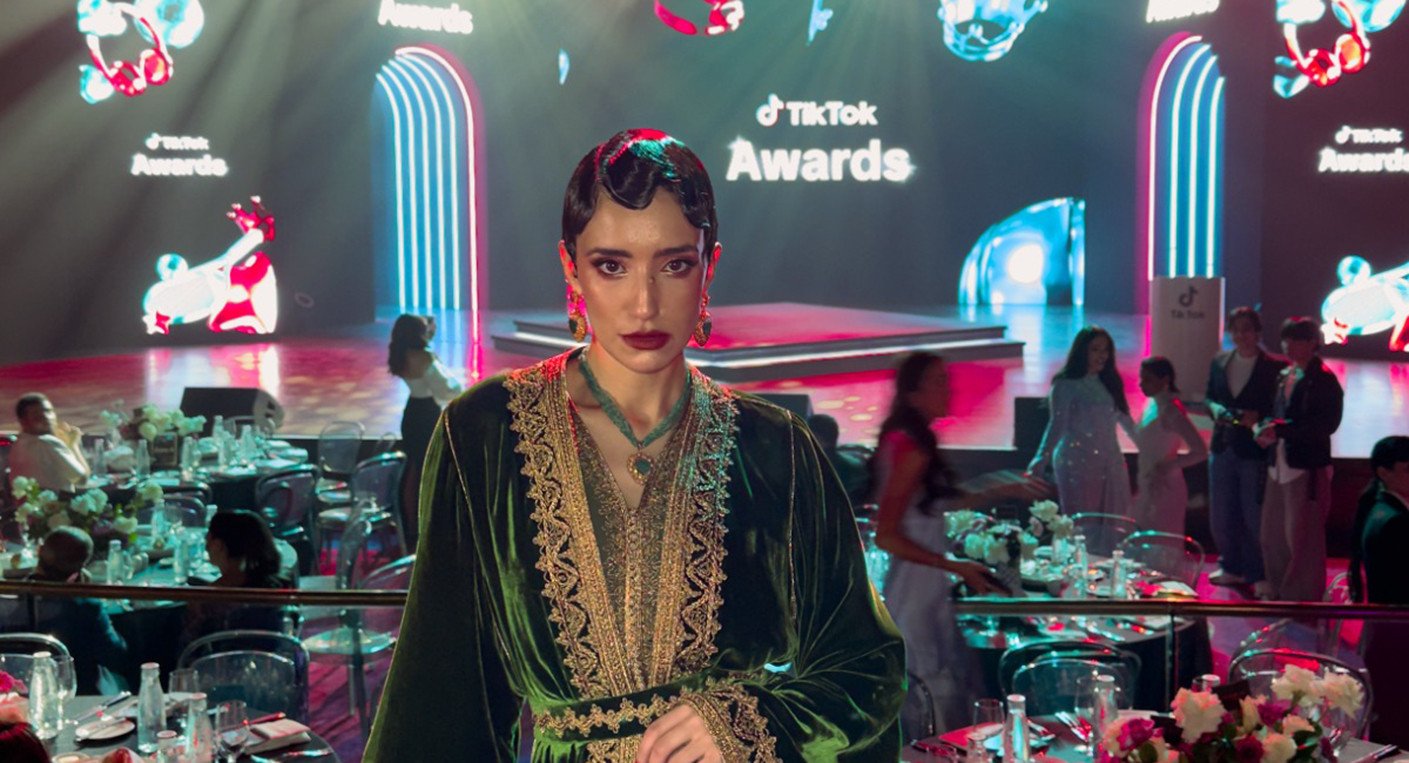 Une jeune femme marocaine souriante, debout devant un paysage urbain, avec un téléphone portable à la main.