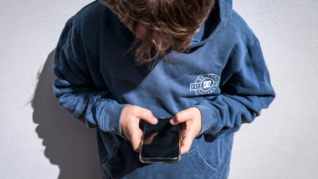 صورة لشاب يلعب على هاتفه المحمول فيما يظهر خلفية لمنصة تواصل اجتماعي.