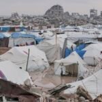 صورة لشاب فلسطيني يرتدي ثوباً يغمره الماء, مع خلفية لخيام نازحين في غزة.