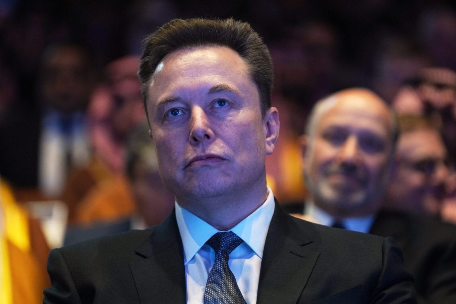 A photo of Elon Musk speaking at a conference, with a serious expression.