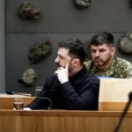 Un groupe de personnes regroupées autour d'un drapeau ukrainien, avec un texte alt "Guerre en Ukraine : Zelensky arrive au Canada pour une rencontre avec le premier ministre"