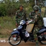 Un soldat thaïlandais se tient debout près d'une voiture de combat, avec un paysage de jungle en arrière-plan.