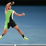 Tennis player in action on a court during the Australian Open, with the stadium visible in the background.