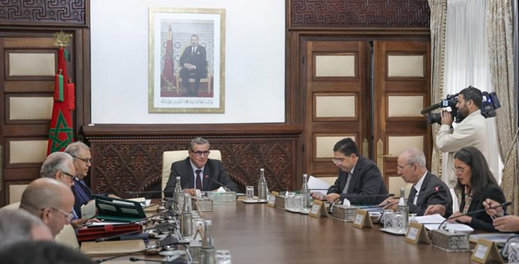 Le Chef du gouvernement Aziz Akhannouch présidant une réunion du Conseil de gouvernement, avec des documents et des ministres en arrière-plan, symbolisant les décisions et réformes à venir.