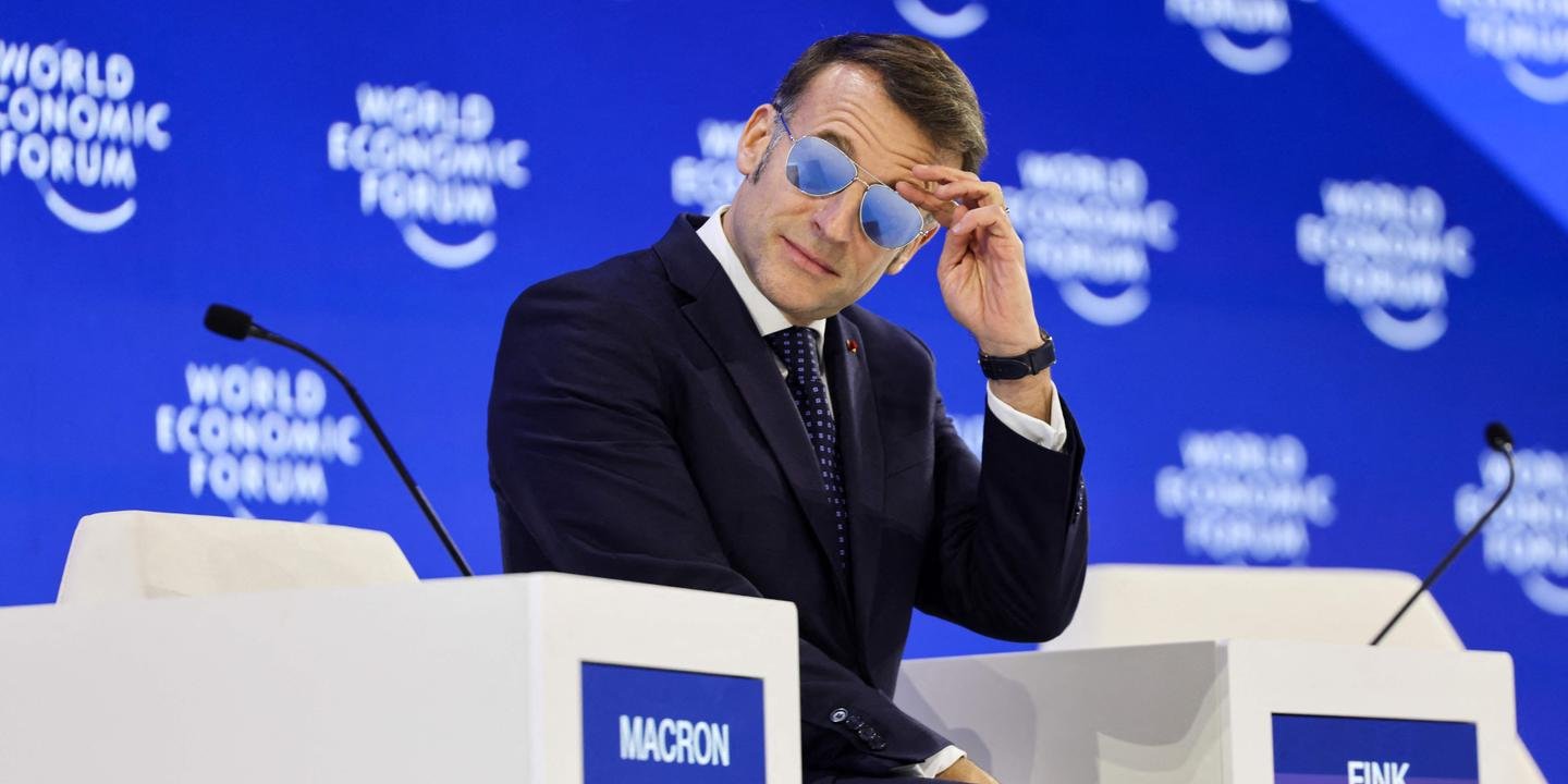 Le président français Emmanuel Macron s'exprimant lors du Forum économique mondial de Davos, avec des drapeaux européens et américains en arrière-plan, symbolisant les tensions évoquées.