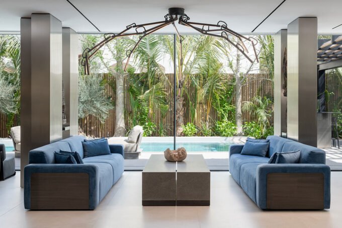 Al Mahra Residence: A Masterclass in Materiality and Dubai Living 9 A contemporary luxury villa interior in Al Mahra, Dubai, showcasing an open-plan living space with brushed stainless steel columns, dark wood, and a striking backlit Patagonia stone bar.