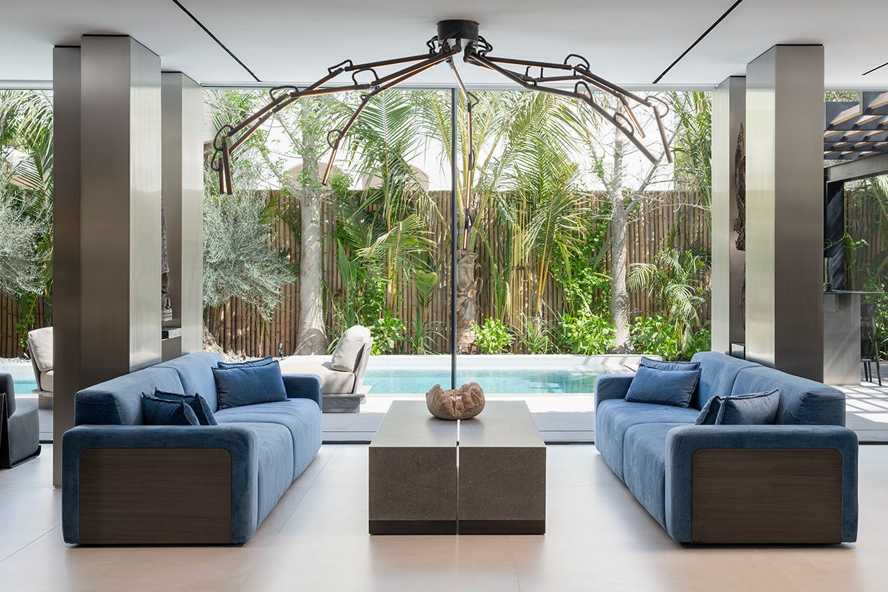 A contemporary luxury villa interior in Al Mahra, Dubai, showcasing an open-plan living space with brushed stainless steel columns, dark wood, and a striking backlit Patagonia stone bar.