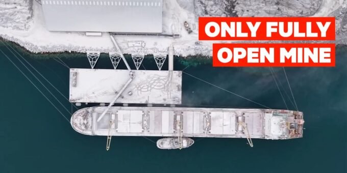 An aerial view of a remote mining operation in the icy, mountainous landscape of Greenland, with heavy machinery visible against the snow.
