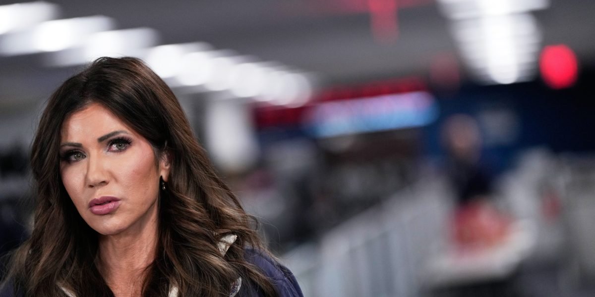 Senator Jacky Rosen speaking at a podium, with a blurred image of Homeland Security Secretary Kristi Noem in the background.