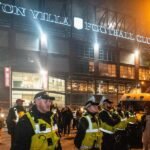 A football stadium with a blurred image of a police officer and a Microsoft Copilot logo overlayed, symbolizing the AI-blamed fan ban controversy.