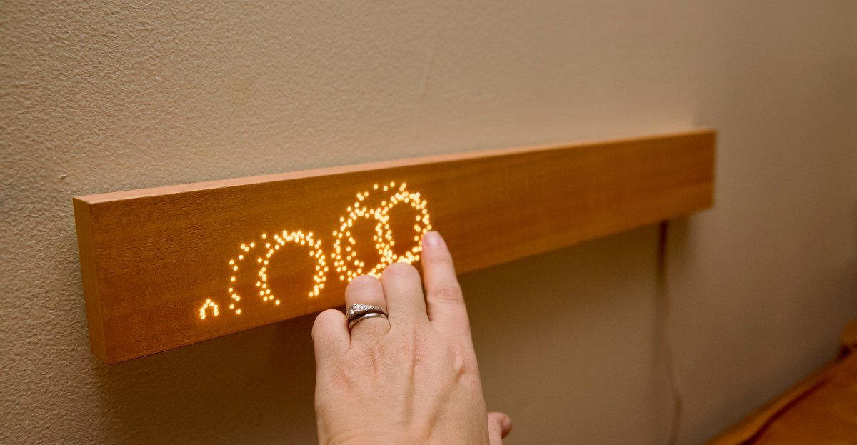 The Mui Board, a minimalist wooden smart home controller, mounted on a wall with glowing dot-matrix icons.