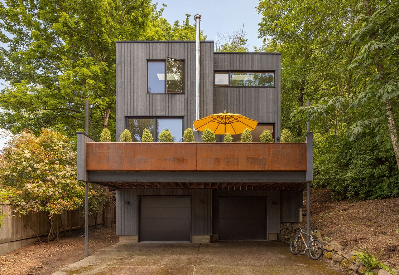 The renovated Alder House exterior, featuring deep navy cedar siding, weathering steel accents, and an elevated deck surrounded by lush Pacific Northwest trees.