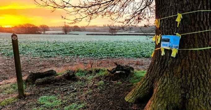Residents gather near an ancient oak tree in a green field, holding a "NO TO DATA CENTRE" protest sign, with a modern data centre rendering in the background.