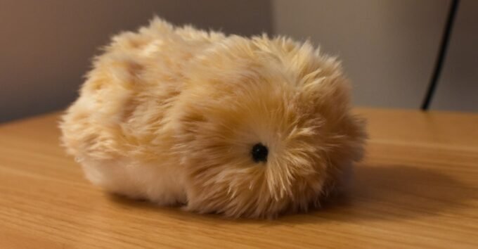Casio Moflin AI pet, a fuzzy, guinea-pig-like robot, sitting on a surface, with a mechanical whirring sound implied.