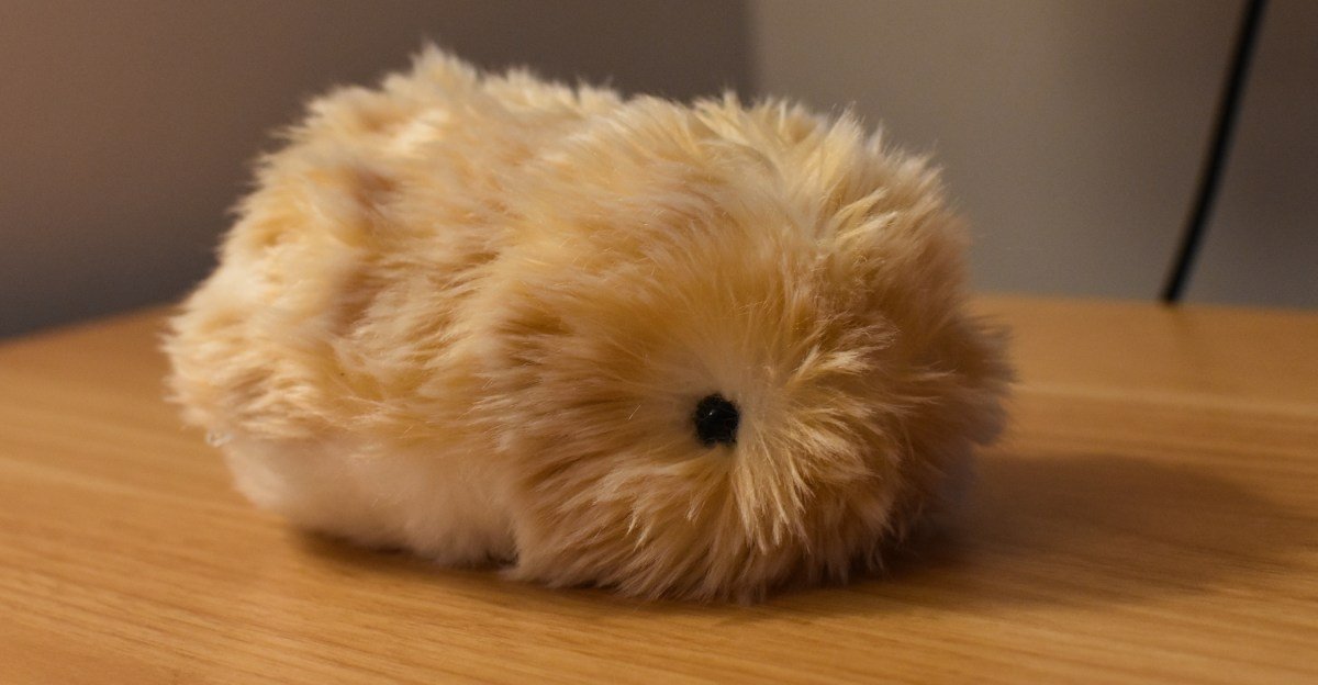 Casio Moflin AI pet, a fuzzy, guinea-pig-like robot, sitting on a surface, with a mechanical whirring sound implied.