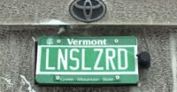 The Lens Lizard module discreetly installed behind a car's rear license plate, showing its nozzle and self-contained design.