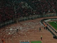 Vue panoramique du Stade international du Caire, avec des débris de bouteilles sur le terrain, un logo de la CAF et une silhouette floue d'un entraîneur, symbolisant les défis du football marocain.
