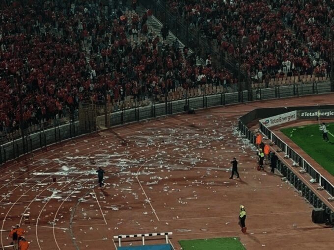 Vue panoramique du Stade international du Caire, avec des débris de bouteilles sur le terrain, un logo de la CAF et une silhouette floue d'un entraîneur, symbolisant les défis du football marocain.