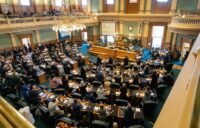 A 3D printer in the process of manufacturing a firearm component, with a blurred background of legislative documents.
