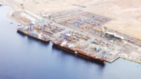 صورة جوية لميناء العقبة الأردني تظهر حركة السفن والحاويات، مع خلفية للمدينة.