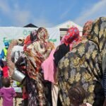 صورة تظهر قافلة مساعدات إنسانية في منطقة متضررة من الصراع في السودان، مع خلفية تعكس الدمار.