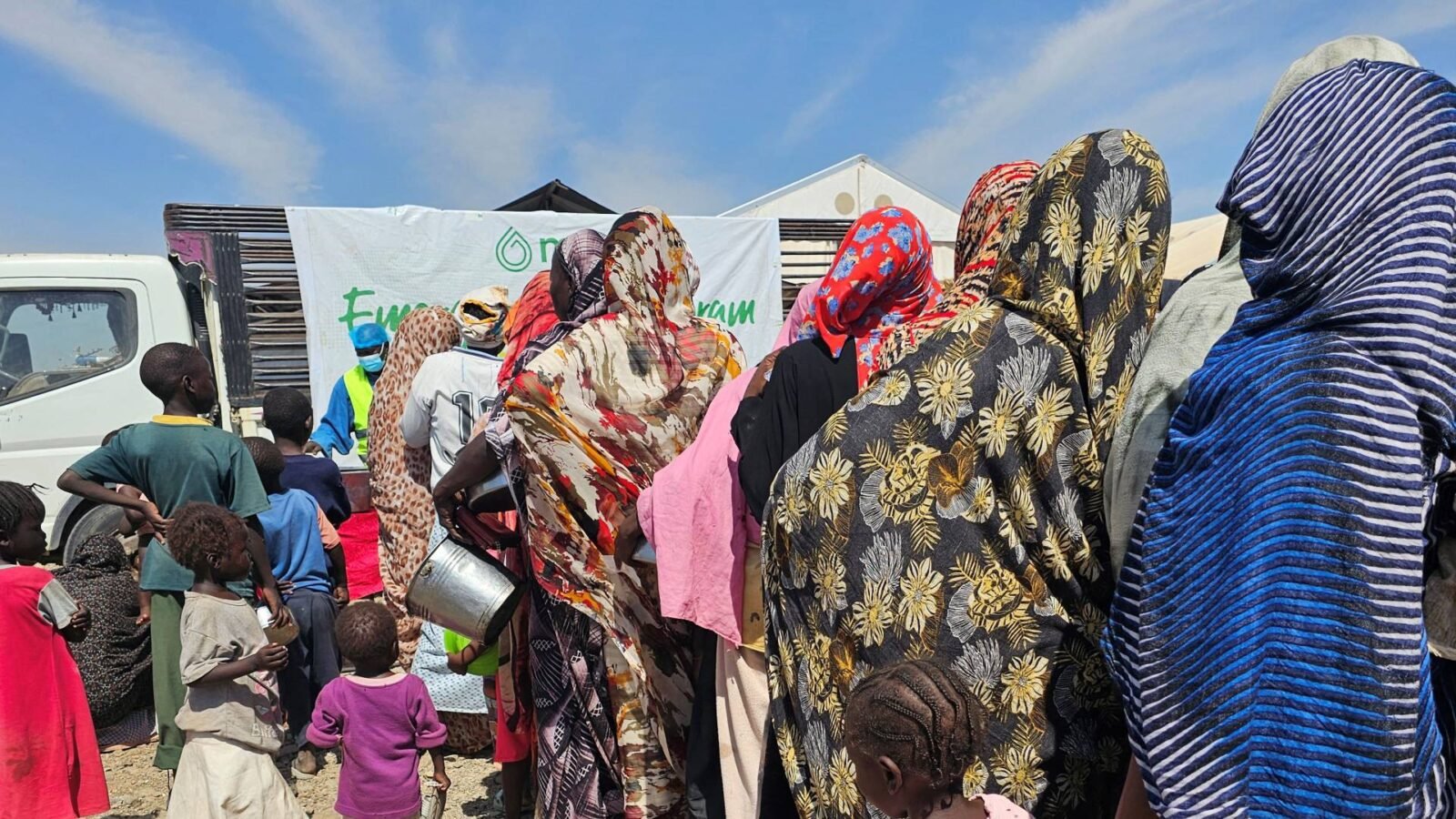 صورة تظهر قافلة مساعدات إنسانية في منطقة متضررة من الصراع في السودان، مع خلفية تعكس الدمار.