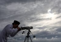 صورة لهلال رمضان يظهر في سماء صافية، يرمز لبداية الشهر الفضيل، مع إضاءة خافتة لمسجد في الأفق.