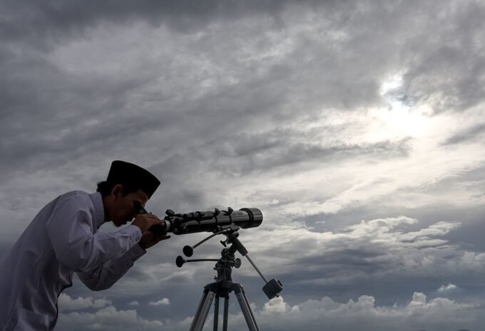 صورة لهلال رمضان يظهر في سماء صافية، يرمز لبداية الشهر الفضيل، مع إضاءة خافتة لمسجد في الأفق.