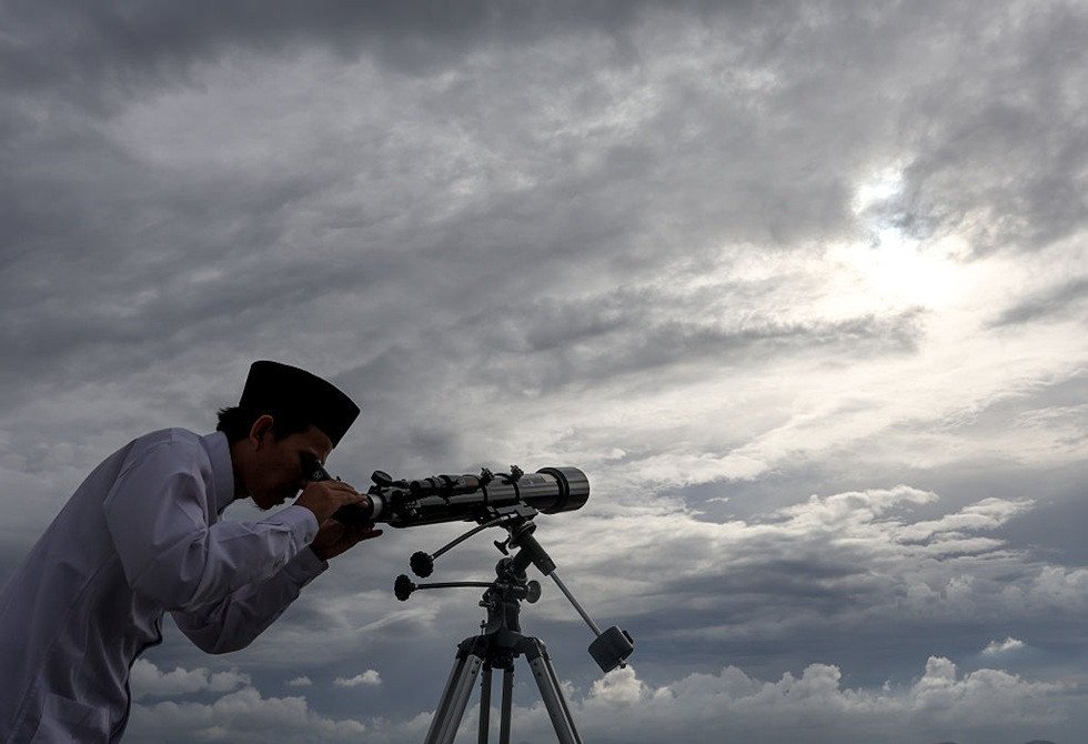 صورة لهلال رمضان يظهر في سماء صافية، يرمز لبداية الشهر الفضيل، مع إضاءة خافتة لمسجد في الأفق.