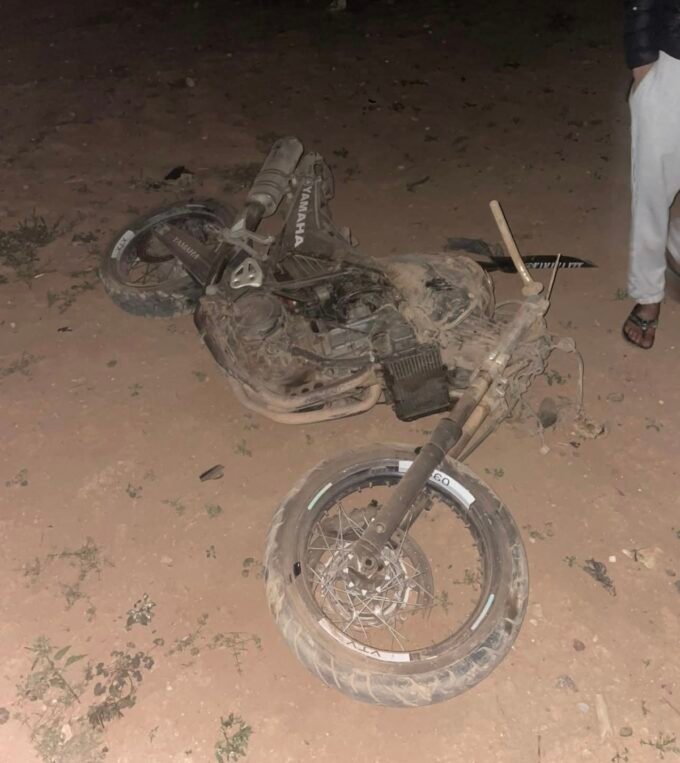 صورة لدراجة نارية متضررة في موقع حادث سير بمدار دائري في أولاد تايمة، مع وجود سيارات الإسعاف والشرطة.