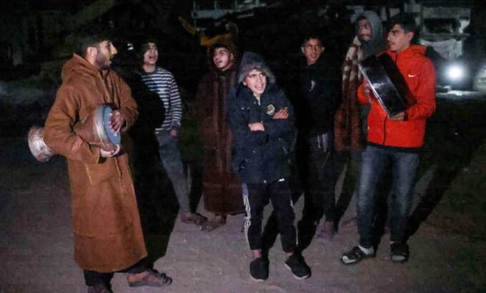 صورة تظهر مسحراتي يقرع الطبل بين خيام النازحين في غزة خلال شهر رمضان، مع أضواء خافتة وأطفال يراقبون.