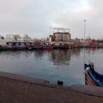 Vue aérienne du port de pêche de Casablanca, montrant des bateaux amarrés et les infrastructures portuaires sous un ciel clair.