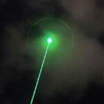 An anti-drone laser system aiming at a drone in the sky over a city skyline.