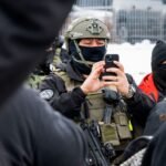 A smartphone screen displaying a facial recognition scan, overlaid with text indicating 'Mobile Fortify' and a blurred image of a person's face, symbolizing the controversial app used by immigration agents.