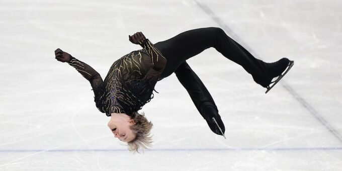 Ilia Malinin mid-air performing a quadruple jump on the ice, surrounded by a blurred audience.