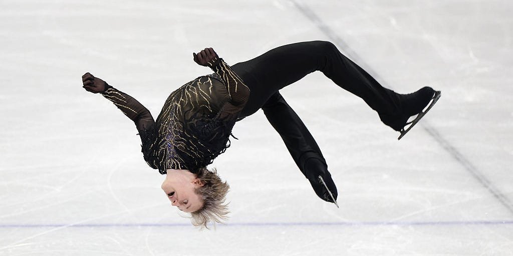 Ilia Malinin mid-air performing a quadruple jump on the ice, surrounded by a blurred audience.