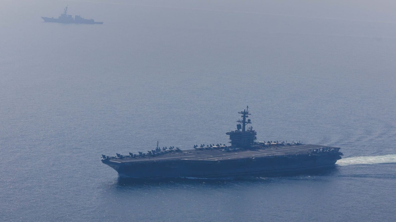 An aerial view of the USS Abraham Lincoln Carrier Strike Group operating in the Arabian Sea, escorted by replenishment ships and Coast Guard vessels, with fighter jets conducting flight operations.