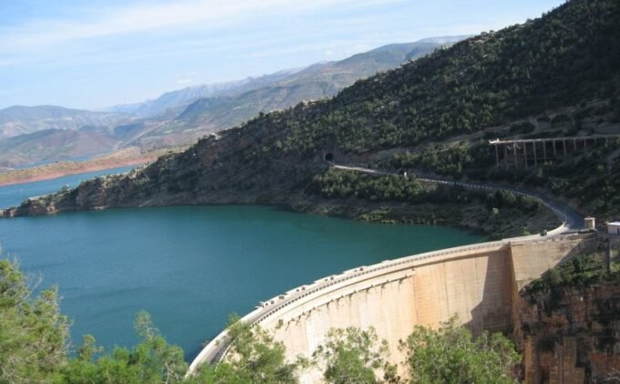 Vue aérienne d'un grand barrage marocain avec un niveau d'eau élevé, entouré de montagnes verdoyantes et de paysages luxuriants, symbolisant l'abondance hydrique retrouvée.