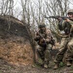 A soldier in muddy combat gear in a trench, with a drone visible in the sky above.