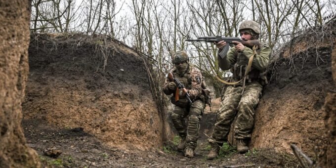 A soldier in muddy combat gear in a trench, with a drone visible in the sky above.