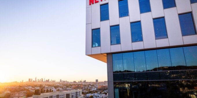 Illustration of Netflix and Warner Bros. logos with a Justice Department scale of justice, symbolizing an antitrust investigation.