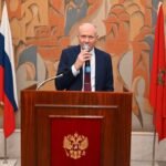 L'ambassadeur de Russie au Maroc, Vladimir Baybakov, s'exprimant lors d'un événement diplomatique, avec un drapeau marocain et russe en arrière-plan.