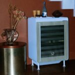 Rocco smart fridge in Ice Blue with fluted glass door, slender steel legs, and an accompanying tray, set in a modern living space.