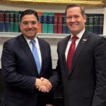 Représentation symbolique des drapeaux des États-Unis, de l'ONU, du Maroc, du Front Polisario, de l'Algérie et de la Mauritanie autour d'une table de négociation, illustrant les pourparlers à Madrid sur le Sahara.