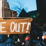 A person holding a smartphone, capturing or viewing an event, with a subtle background suggesting conflict or observation.