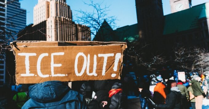 A person holding a smartphone, capturing or viewing an event, with a subtle background suggesting conflict or observation.