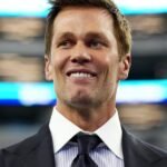 Tom Brady in a broadcast booth, wearing a suit and headset, speaking into a microphone with a focused expression.