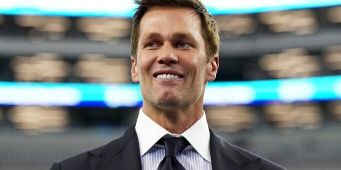 Tom Brady in a broadcast booth, wearing a suit and headset, speaking into a microphone with a focused expression.