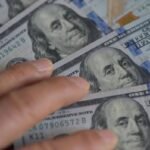 President Donald Trump speaking at an economic event, with financial charts or currency symbols in the background.