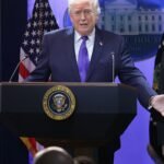Donald Trump speaking at a podium, with a backdrop of American flags, discussing trade policy.